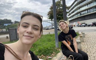 Two people smiling near a street in Porto with a modern building and bridge in the background.