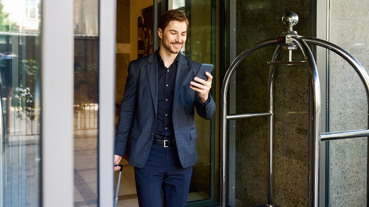 Businessman with suitcase checking smartphone outside hotel.