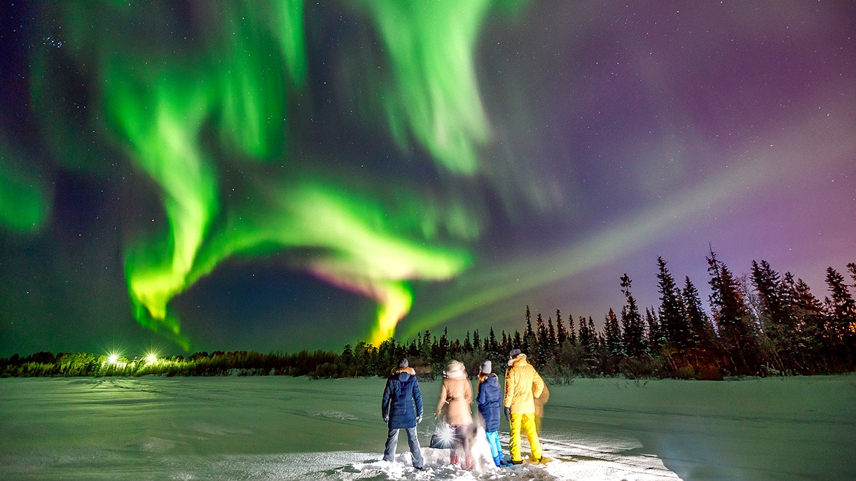 Group of tourists on a Northern Lights tour in a snowy landscape at night, gazing at the vibrant aurora borealis in the sky