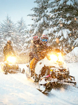Snowmobile riders navigating snowy forest trail.