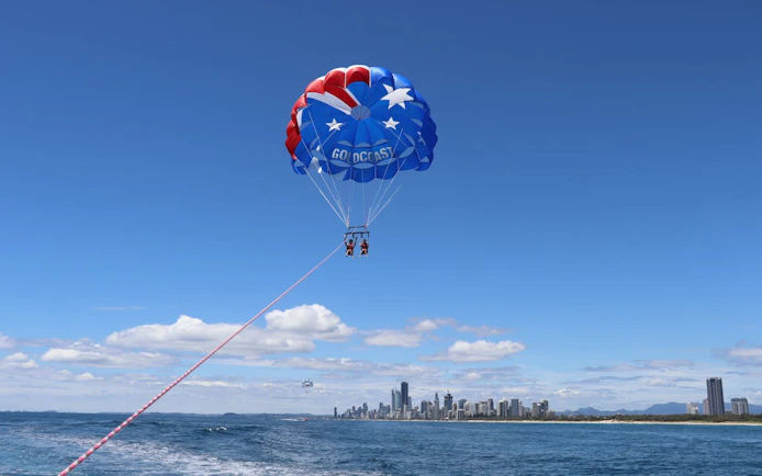 Three people parasailing over Gold Coast waters with city skyline in background.
