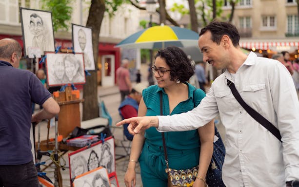 Couple exploring street artist's caricatures in Montmartre, Paris during guided walking tour.