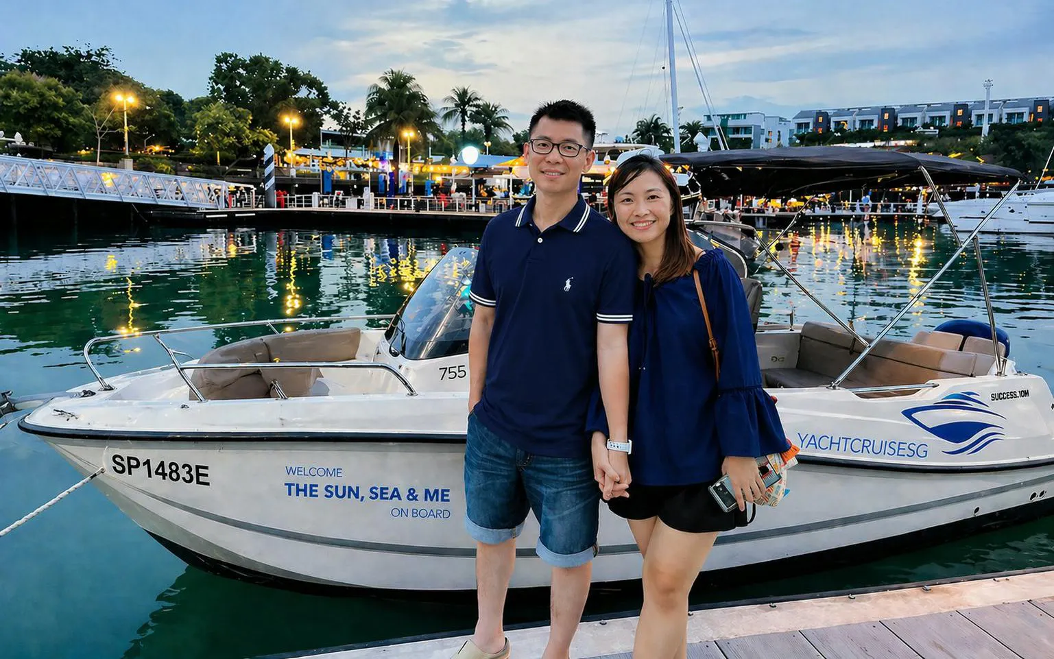Couple standing by YachtCruiseSG speedboat at marina, Singapore evening lights reflecting on water.