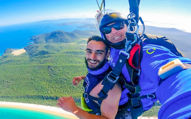 Tandem skydivers over Airlie Beach, showcasing turquoise waters and lush landscape below.