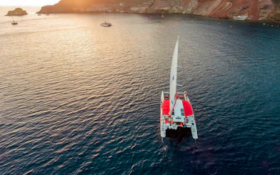 Catamaran sailing at sunset near Santorini cliffs.
