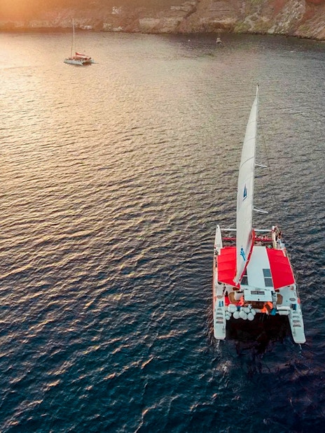 Catamaran sailing at sunset near Santorini cliffs.