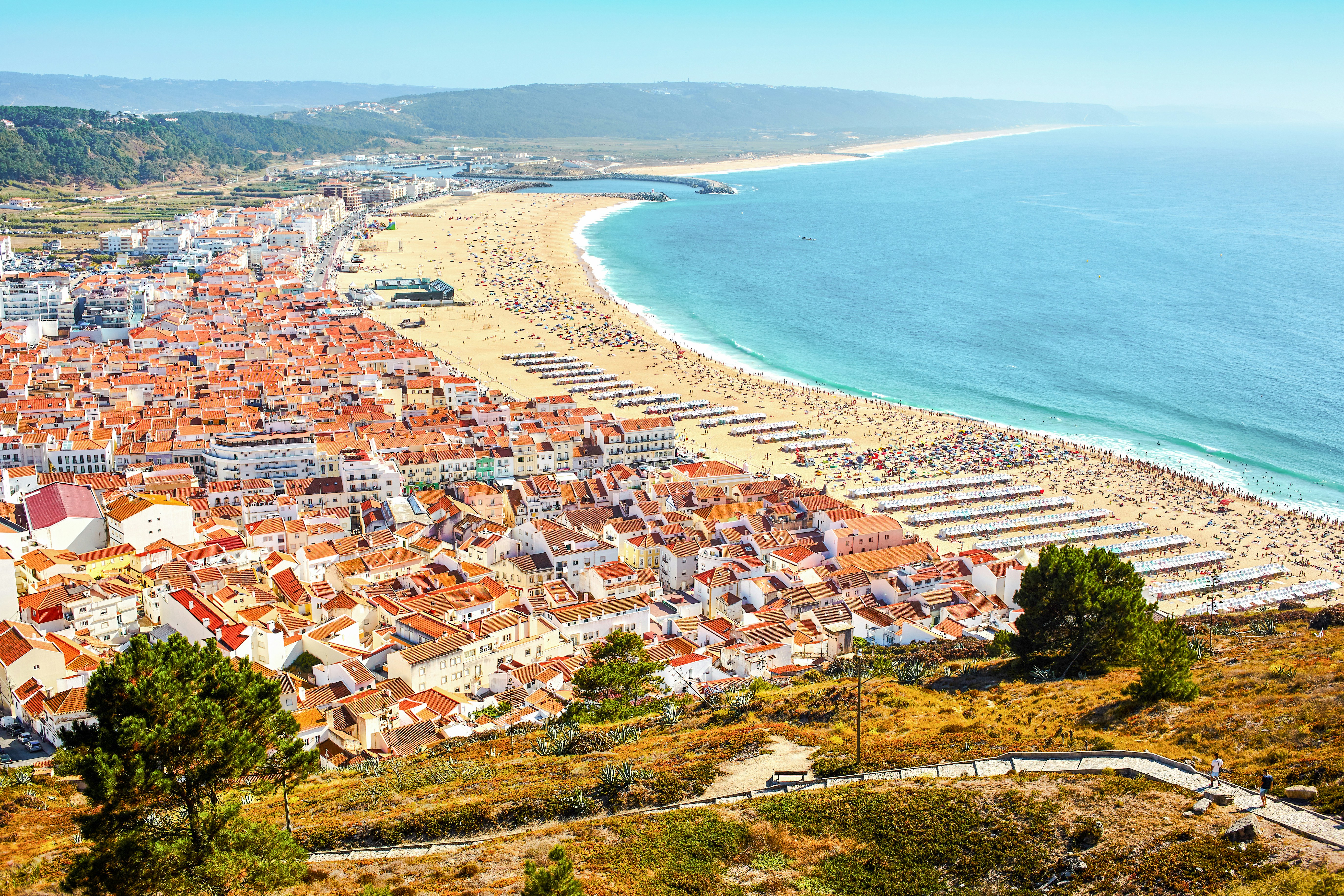 Aerial view of Nazare city, Portugal, showing red-roofed buildings and a crowded beach.