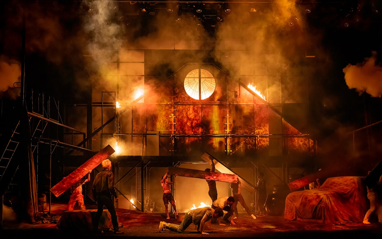 Stage scene from "The Outsiders" with actors amidst a fiery backdrop.
