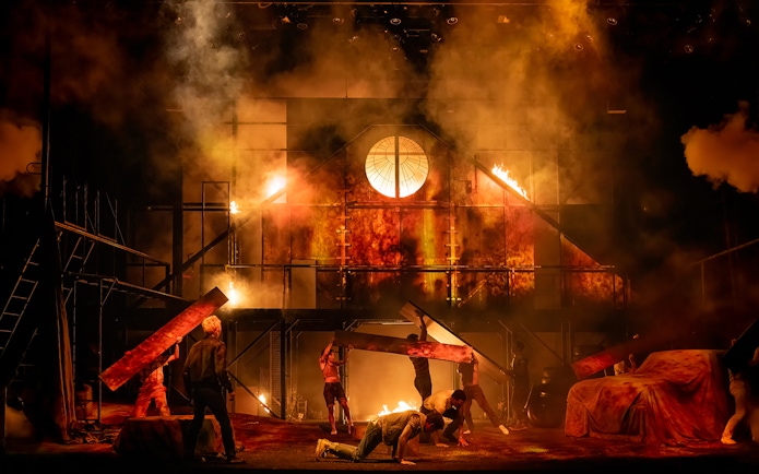Stage scene from "The Outsiders" with actors amidst a fiery backdrop.