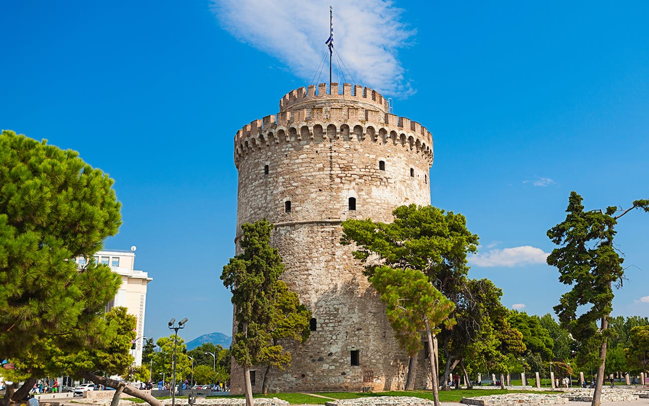 White Tower of Thessaloniki surrounded by trees under a clear blue sky.