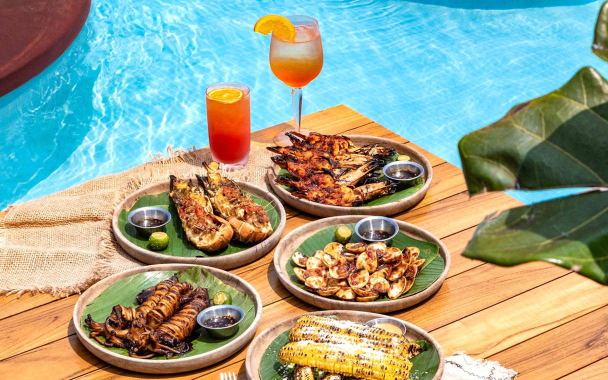 Grilled seafood and tropical drinks by a pool, part of Food Discovery Pass experience.