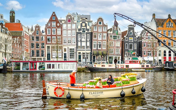 Guests enjoying tulip cruise on Amsterdam canal with historic buildings in background.