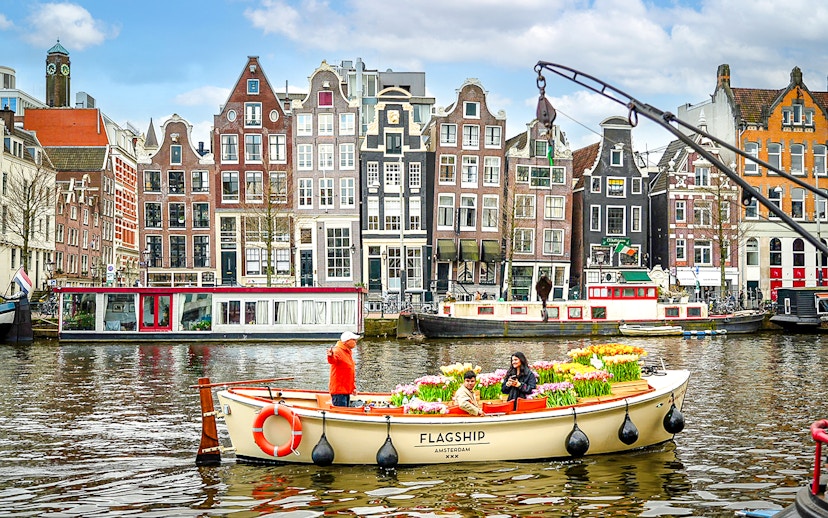 Guests enjoying tulip cruise on Amsterdam canal with historic buildings in background.
