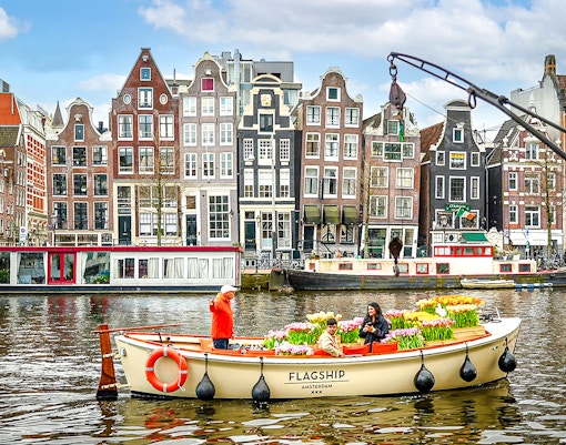 Guests enjoying tulip cruise on Amsterdam canal with historic buildings in background.