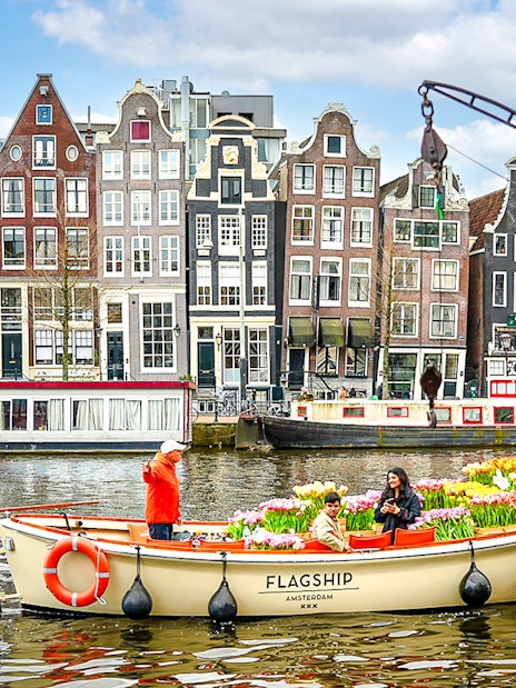 Guests enjoying tulip cruise on Amsterdam canal with historic buildings in background.