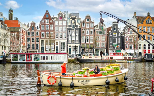 Guests enjoying tulip cruise on Amsterdam canal with historic buildings in background.