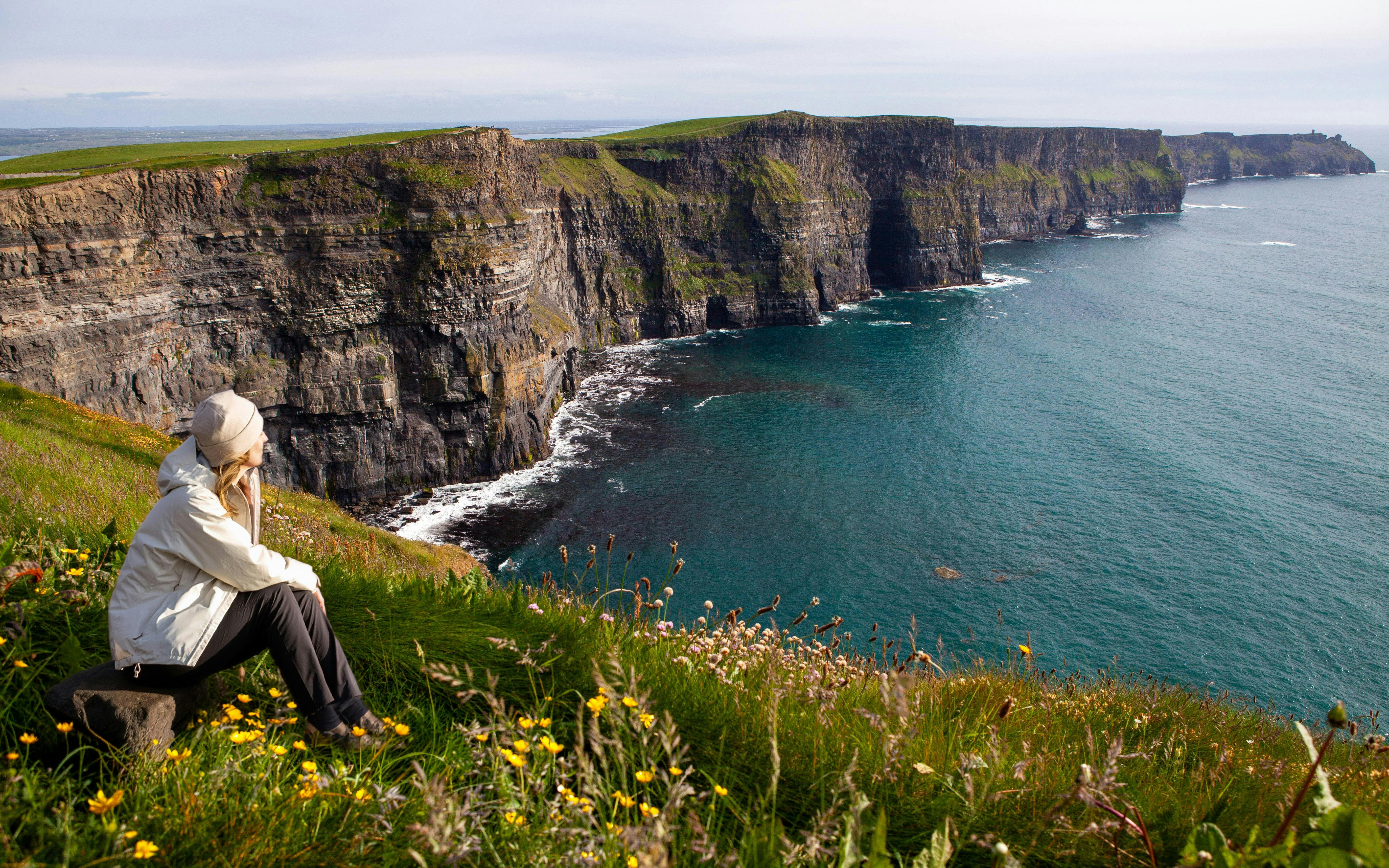 Person sitting on grassy cliff edge overlooking Cliffs of Moher and Atlantic Ocean in Ireland.