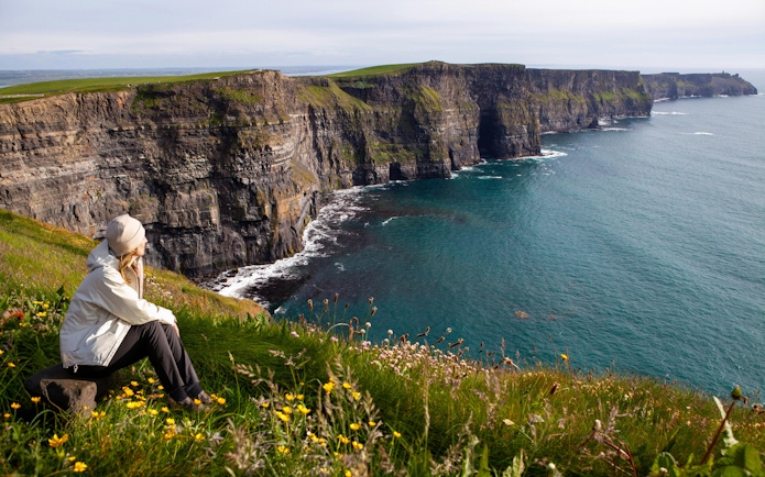 Person sitting on grassy cliff edge overlooking Cliffs of Moher and Atlantic Ocean in Ireland.