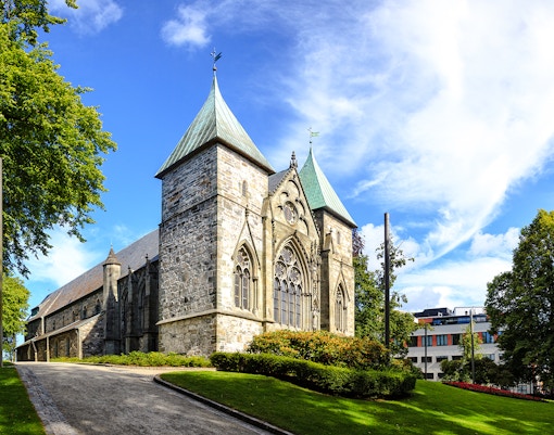 Stavanger Cathedral with stone facade and green spires surrounded by trees and a clear sky.