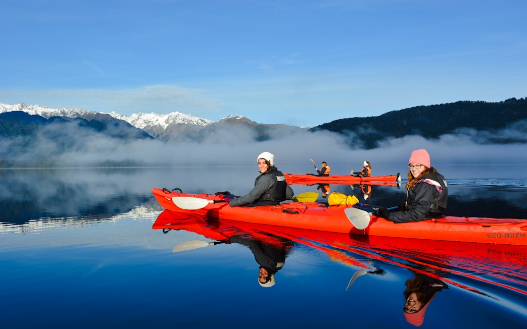 Visitors kayaking on Lake Mapourika with misty mountains in the background.