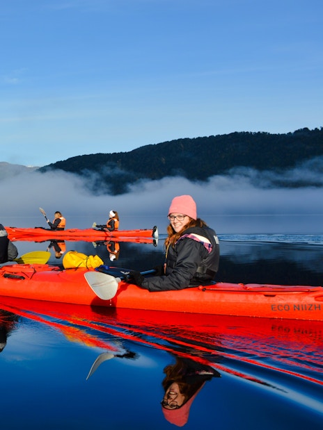 Visitors kayaking on Lake Mapourika with misty mountains in the background.