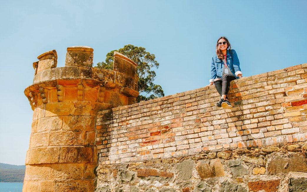 Person sitting on historic stone wall at Port Arthur, Tasmania, with scenic view in background.