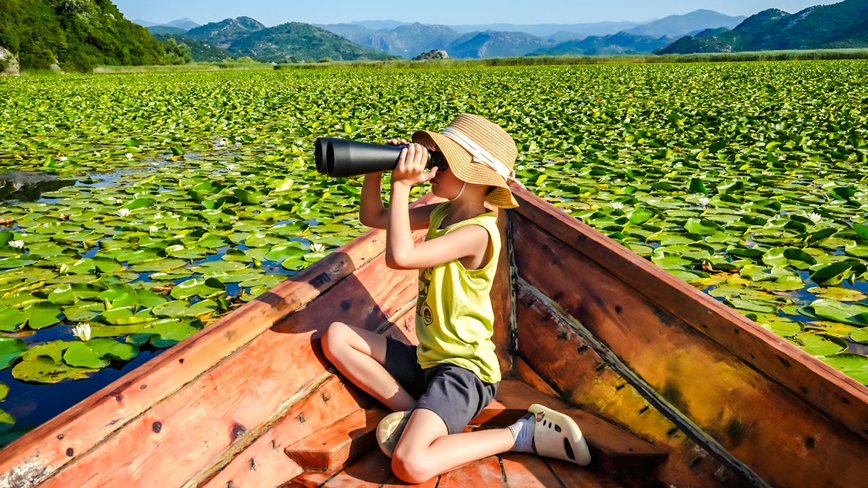 Boy with binoculars on boat at Skadar Lake, Montenegro, surrounded by lily pads.