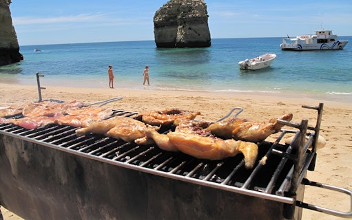 Grilled chicken on a beach with boats and rock formations in Benagil, Portugal.