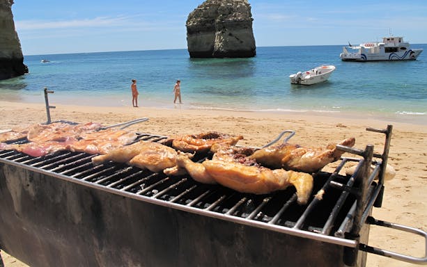 Grilled chicken on a beach with boats and rock formations in Benagil, Portugal.