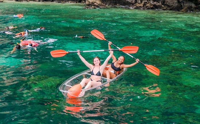 Two women in a clear kayak with paddles, surrounded by snorkelers in emerald water.