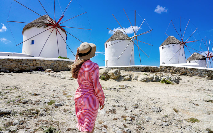 Woman in a hat walking towards iconic windmills on Mykonos, Greece.
