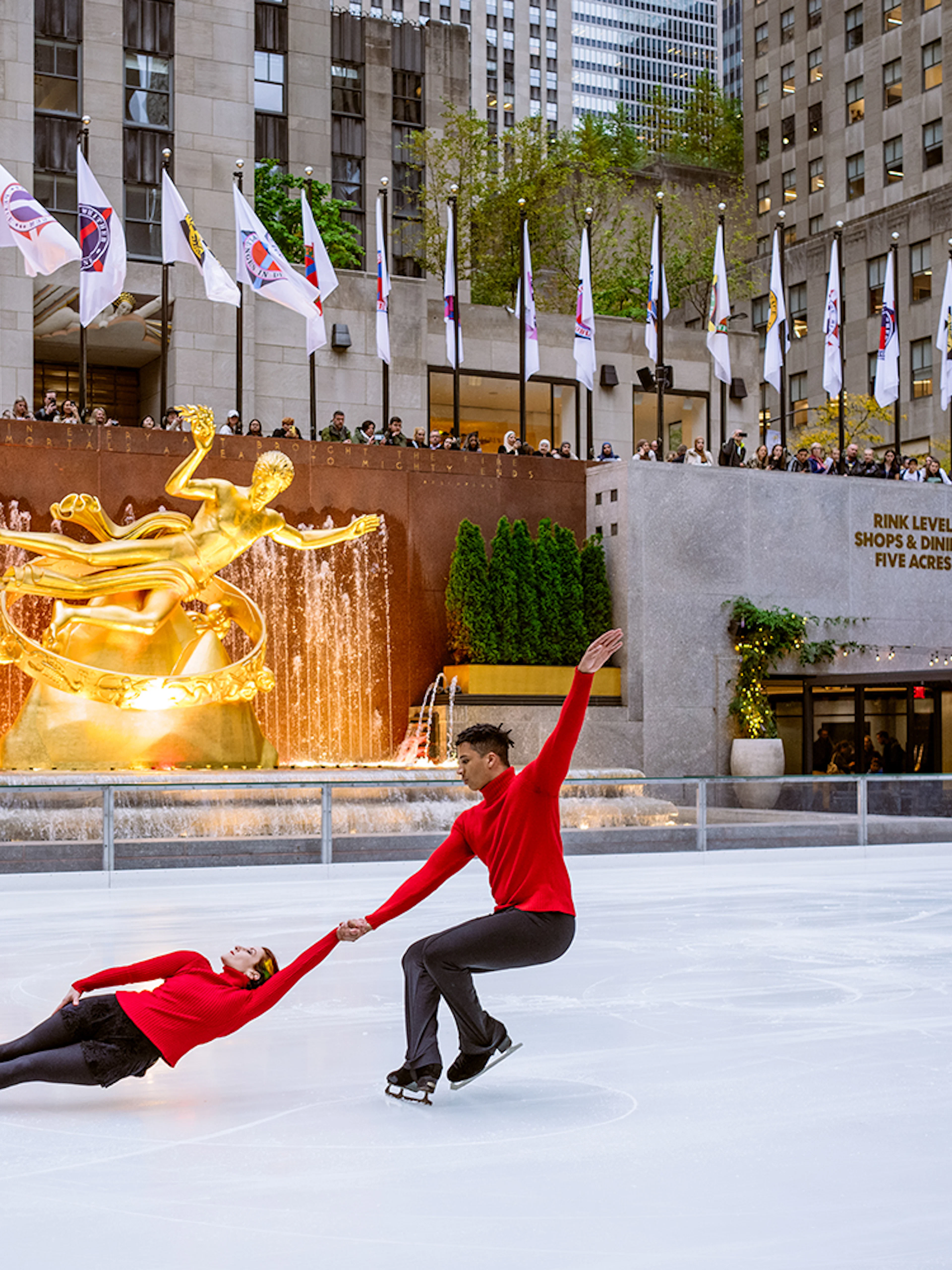 Ice skaters performing at Rockefeller Center's The Rink with Prometheus statue in the background.