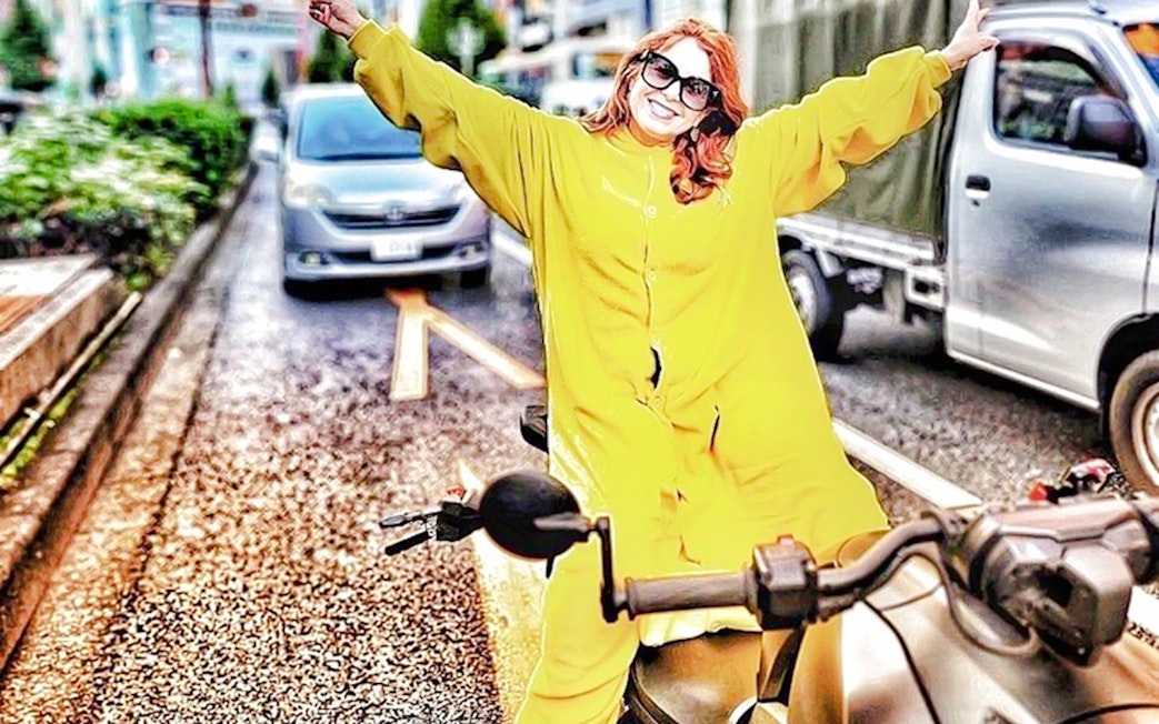 Woman in yellow onesie on go-kart with arms outstretched on Tokyo street.