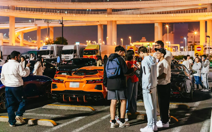 Group gathers near sports cars at Tokyo parking lot, part of Fast & Furious Tokyo Drift III Experience.