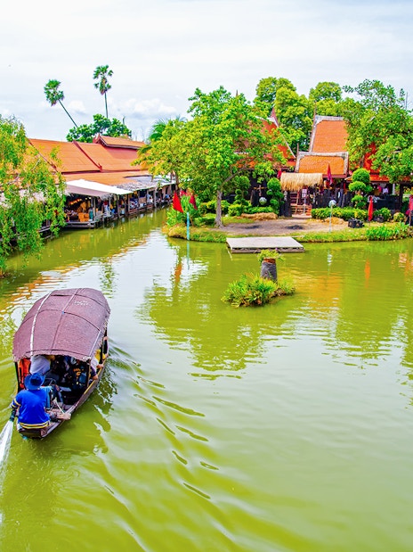 Boat navigating Ayutthaya Floating Market canal with traditional buildings and lush greenery.