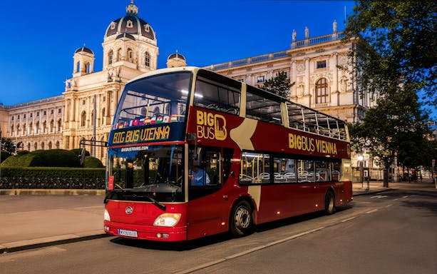 Red double-decker bus on Vienna evening tour passing historic building.