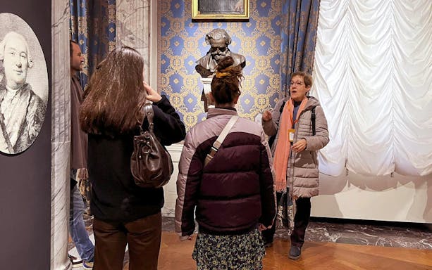 Guide explaining to tourists inside La Scala Theatre with a bust in the background.