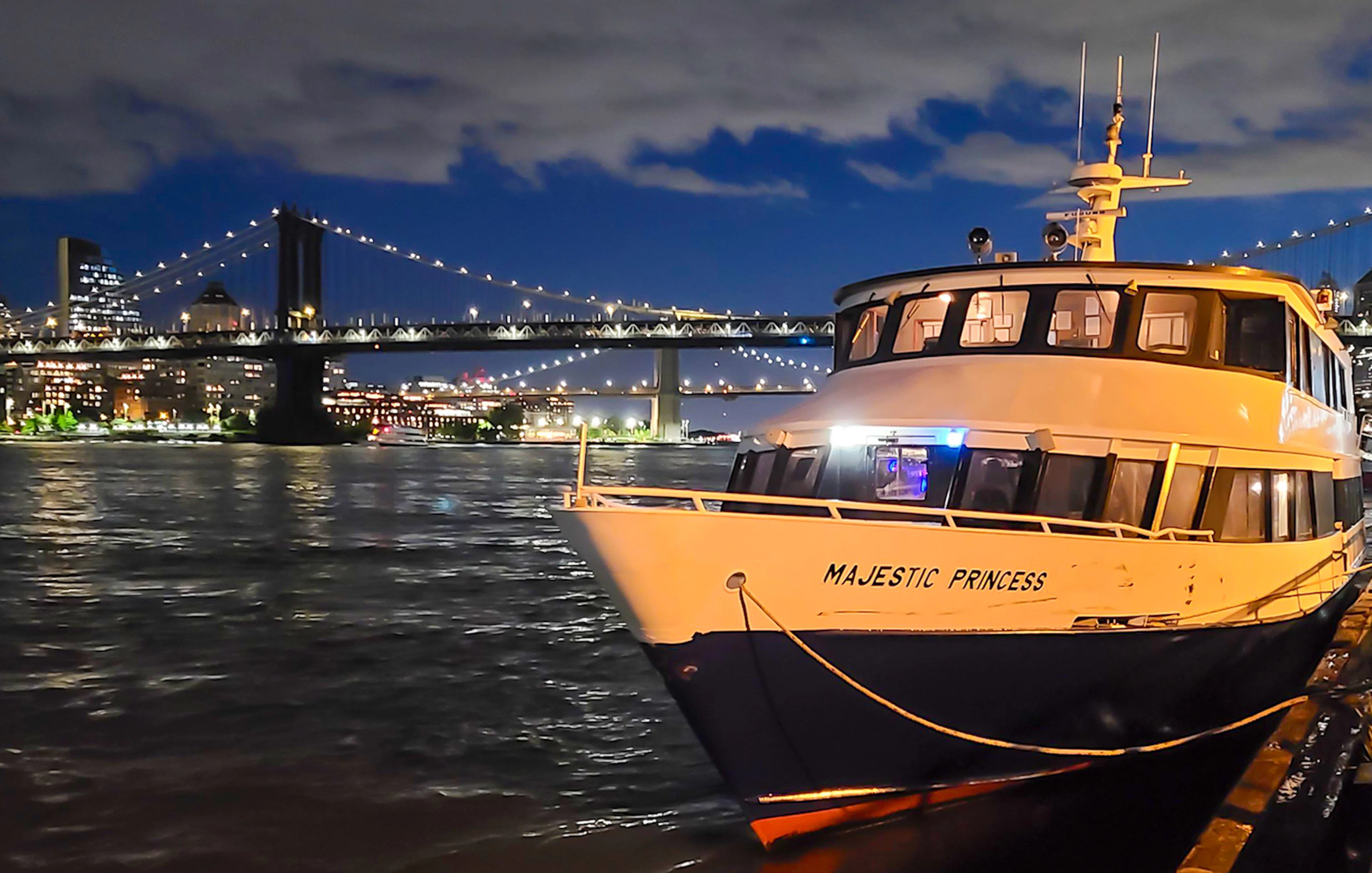 Cruise boat docked with NYC skyline and bridge illuminated at night.