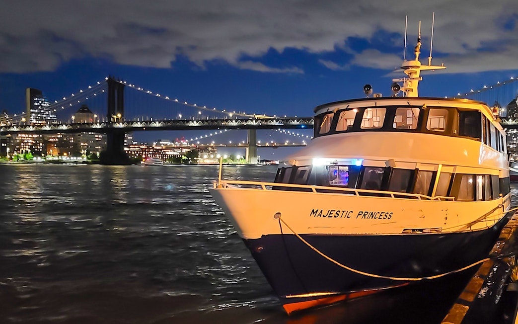 Cruise boat docked with NYC skyline and bridge illuminated at night.
