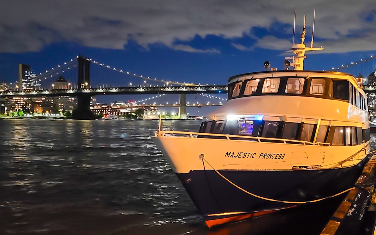 Cruise boat docked with NYC skyline and bridge illuminated at night.