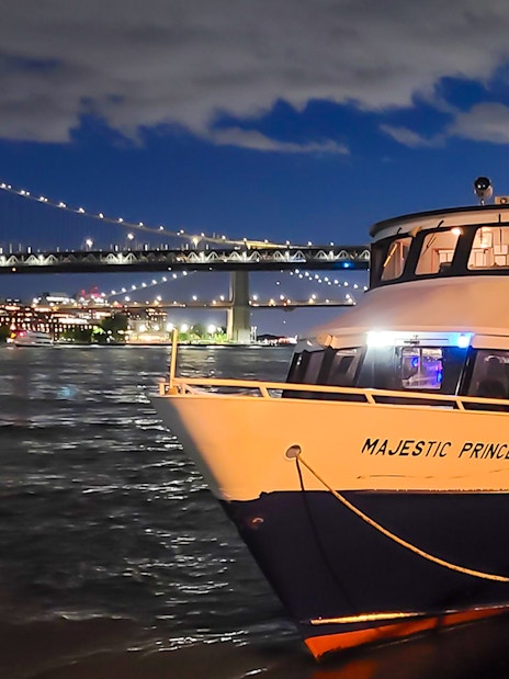 Cruise boat docked with NYC skyline and bridge illuminated at night.