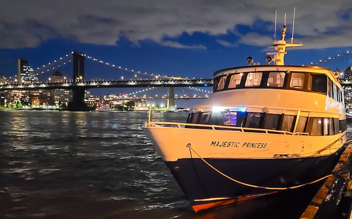 Cruise boat docked with NYC skyline and bridge illuminated at night.