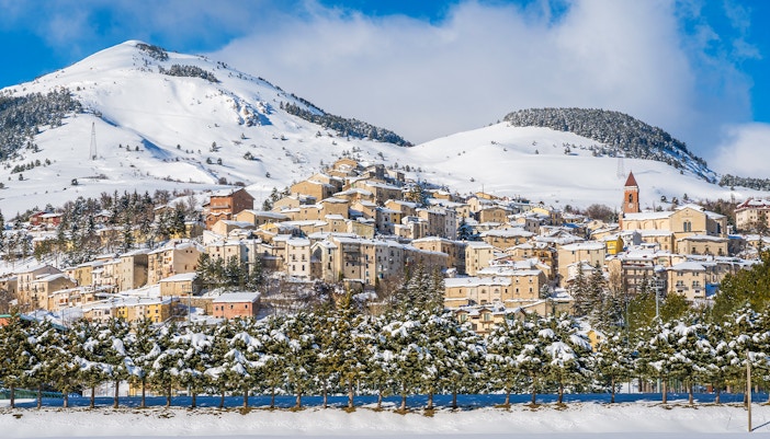 Skiers on snowy slopes in Roccaraso, Italy, with pine trees and mountains in the background.