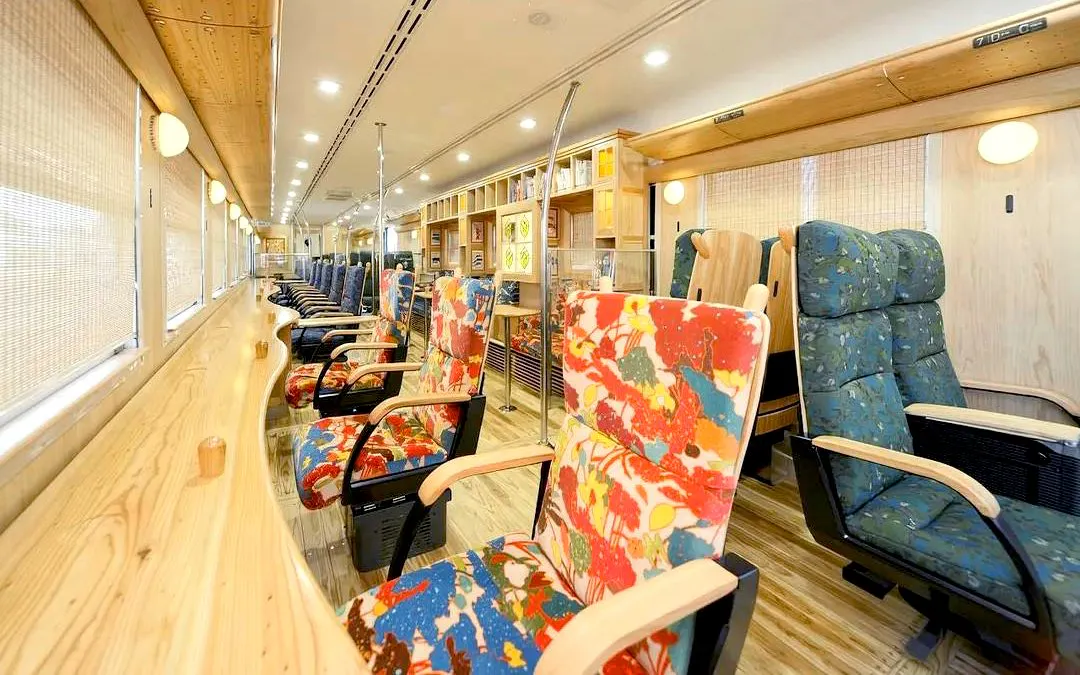 Colorful seating area inside JR Kyushu train with wooden interior and large windows.