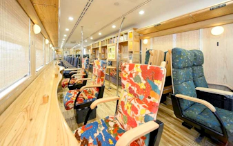 Colorful seating area inside JR Kyushu train with wooden interior and large windows.