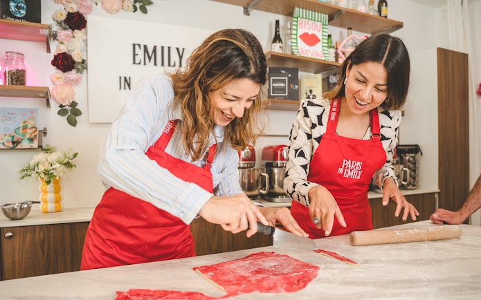 People making croissants at Emily in Paris workshop, using red dough and kitchen tools.