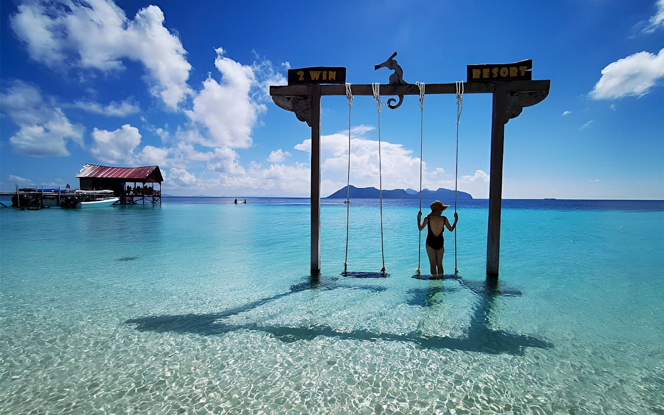 Person on swing over clear water at Mataking Island, Semporna, with distant islands and a dock.