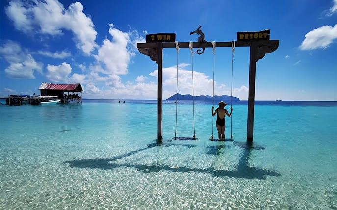 Person on swing over clear water at Mataking Island, Semporna, with distant islands and a dock.