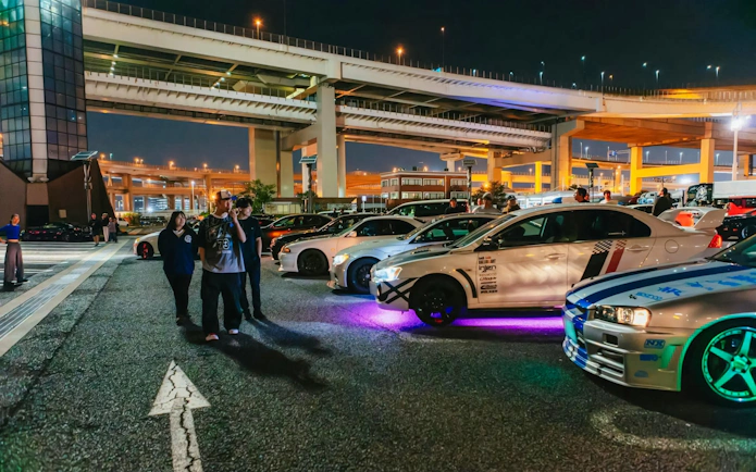 Cars and people at a Tokyo parking lot under highway lights, part of the Fast & Furious Tokyo Drift III Experience.