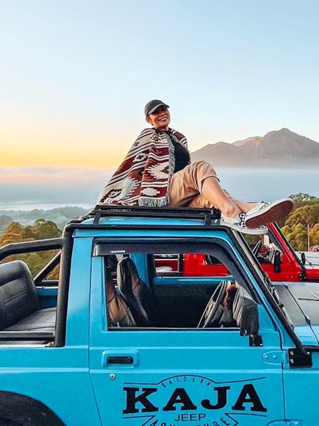 Person sitting on a blue jeep with Mount Batur in the background at sunrise, Bali.